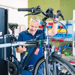 Lächelnder Fahrradmechaniker im blauen Hemd arbeitet an einem blauen E-Bike in einer professionellen Werkstatt mit Werkzeugen an der Wand.