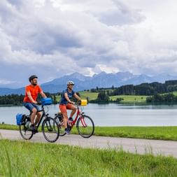 Zwei Radfahrer fahren am Forggensee entlang mit Bergpanorama. Grüne Wiesen im Vordergrund, Alpengipfel und bewölkter Himmel im Hintergrund.