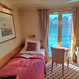 Gemütliche Kabine auf der MS Casanova mit Einzelbett, roter Bettwäsche, Schreibtisch, Stuhl und Glastür mit Flussblick auf historische Gebäude.