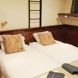 Gemütliche Doppelkabine mit weißer Bettwäsche, beigen Kissen, dunkler Holzvertäfelung, Leiter und Bullauge auf dem Schiff Simmer fan Fryslan.