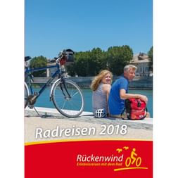 Cover des Rückenwind Radreisen Katalogs 2018 mit Paar am türkisfarbenen Wasser sitzend, Fahrrad, rote Taschen und Helm sichtbar.