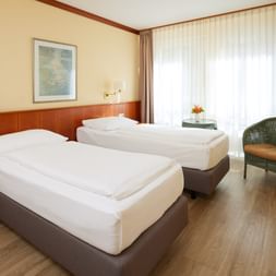 Helles Hotelzimmer mit zwei Einzelbetten, Holzkopfteil, grünem Sessel, Stehlampe und großem Fenster mit Gardinen im Hotel Geheimer Rat.