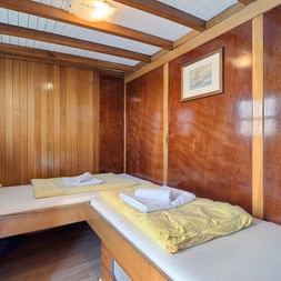 Holzvertäfelte Zweibettkabine auf der MY Linda mit zwei Einzelbetten, gelber Bettwäsche, weißen Handtüchern, Bullauge und Balkendecke.