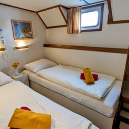 Zweibettkabine mit zwei Einzelbetten, weißer Bettwäsche, gelben Handtüchern, Bullauge, Wandlampen, gerahmten Bildern und Holzregal.