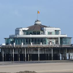 Modernes Seebrückengebäude mit Glasfassade und Kuppel in Blankenberge, Belgien, auf Betonsäulen über Sandstrand mit belgischer Flagge.