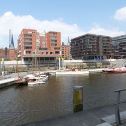 Marina in der HafenCity Hamburg mit Booten an Holzstegen. Moderne Wohngebäude mit roten Backstein- und Glasfassaden säumen das Ufer.