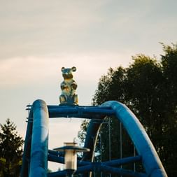 Blaue Bärenfigur sitzt auf blauem Spielplatzgerät mit gebogenen Metallrohren, umgeben von grünen Bäumen unter bewölktem Himmel.