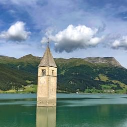 Historischer Kirchturm mit spitzem Dach ragt aus dem türkisfarbenen Wasser des Reschensees, umgeben von grünen Alpenbergen unter bewölktem Himmel.