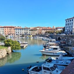 Malerischer Hafenkanal in der Toskana mit weißen Booten an Steinmauern, bunte historische Gebäude säumen das Ufer unter blauem Himmel.