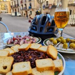 Fahrradhelm und Bierglas auf Tisch mit spanischen Tapas: Oktopus, Oliven und Brot mit dunklem Aufstrich. Straßenszene in Valencia im Hintergrund.