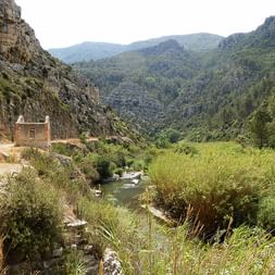 Grünes Flusstal in der Region Valencia mit felsigen Klippen, dichter Vegetation und einem Steingebäude. Berge erheben sich im Hintergrund.