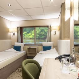 Modern ship cabin on MS Lisabelle with two single beds, green chair, wooden desk, and large windows showing river landscape outside.