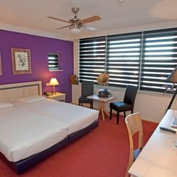 Helles Hotelzimmer mit lila Akzentwand, Doppelbett, rotem Teppich, Schreibtisch, Sitzbereich und großen Fenstern mit Streifenjalousien.