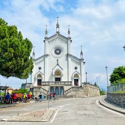 Weiße Kirche Chiesa Parrocchiale di Vendoglio mit Rosette und Glockentürmen. Radfahrer versammeln sich an der Eingangstreppe in Friaul.