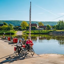 Zwei Radfahrer mit Tourenrädern und Packtaschen auf einer kleinen Fähre über die Weser bei Hemeln, mit Dorfhäusern und Hügeln im Hintergrund.