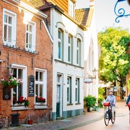 Zwei Radfahrer fahren durch eine malerische Kopfsteinpflasterstraße in Leer, Ostfriesland, gesäumt von bunten historischen Gebäuden.
