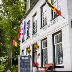 Weiße Fassade des Hans Restaurants in Gaasterland mit deutschen, friesischen und belgischen Flaggen. Eine Kreidetafel steht davor unter grünen Bäumen.