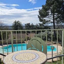 Balcony with mosaic table and chairs overlooking a turquoise pool, surrounded by trees and Provencal landscape under blue sky.
