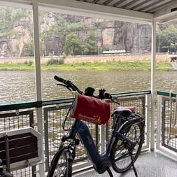 Blaues E-Bike mit roter Rückenwind-Tasche auf Elbfähre. Blick auf Fluss, grünes Ufer und markante Sandsteinfelsen im Hintergrund.