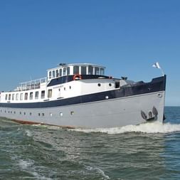 Weiß-dunkelblaues Passagierschiff Floor van Aemstel fährt auf ruhigem Wasser unter klarem blauen Himmel, mit weißer Flagge am Heck.