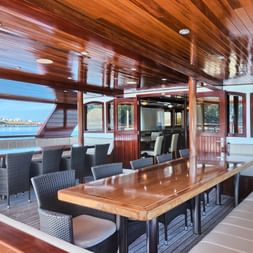 Geräumiges Deck der MY Andela Lora mit Holzdecke, Esstischen, Korbstühlen und gepolsterten Bänken. Große Fenster bieten Küstenblick.
