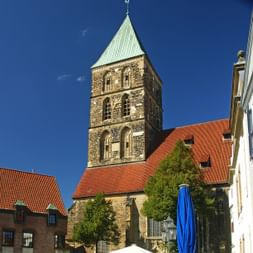 Mittelalterlicher Steinkirchturm mit grüner Kupferspitze in Rheine, umgeben von Gebäuden mit roten Ziegeldächern unter blauem Himmel.