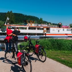 Zwei Radfahrer mit roten Packtaschen stehen bei ihren Rädern auf gepflastertem Platz und beobachten ein weißes Fahrgastschiff auf der Weser.