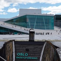 Opernhaus Oslo mit weißem Schrägdach und Glasfassade. Im Vordergrund eine Holzstruktur mit 'Oslo Badstuforening'-Schild am Wasser.