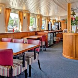 Helles Restaurantinterieur auf der Floor van Aemstel mit Holztischen, bunten Stühlen, geschwungener Bar und großen Fenstern mit orangen Vorhängen.