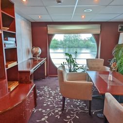 Bibliothekslounge an Bord der SE Manon mit beigen Sesseln, Holzregalen, Pflanzen und großem Panoramafenster mit Blick auf Flusslandschaft.