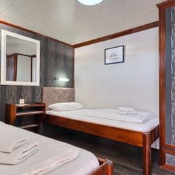 Zweibettkabine auf MY Magellan mit zwei Betten, Holzschrank, Spiegel, Wandlampen und gemusterten Wandpaneelen. Weiße Handtücher auf Betten.