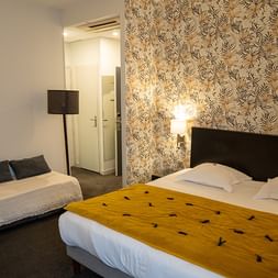 Hotelzimmer im Grand Hotel Toulouse mit zwei Betten, schwarzen Kopfteilen, gelber Decke, Blumentapete, Stehlampe und Eingang zum Bad.