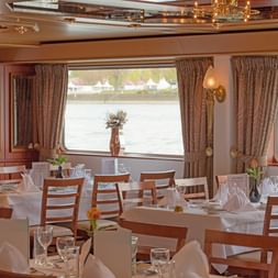 Eleganter Speisesaal auf der MS Switzerland mit gedeckten Tischen, Holzstühlen, weißen Tischdecken und großen Fenstern mit Flussblick.