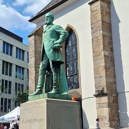 Bronzestatue von Alfred Krupp auf einem Sockel vor einem weißen Gebäude mit Steinsäulen und Rundbogenfenster in Essen.