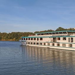 Flusskreuzfahrtschiff MS Switzerland mit weißem Rumpf und grünen Akzenten auf ruhigem Fluss, umgeben von Bäumen unter blauem Himmel.