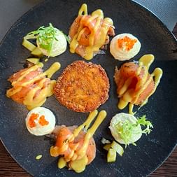 Gourmet-Vorspeise auf schwarzem Schieferplatte mit knusprigem paniertem Plätzchen in der Mitte, umgeben von Räucherlachs und Frischkäse mit Kaviar.
