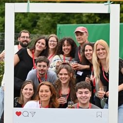 Gruppe von Kollegen posiert in einem großen weißen Instagram-Rahmen bei einer Outdoor-Feier, trägt rote Bänder und hält Getränke.