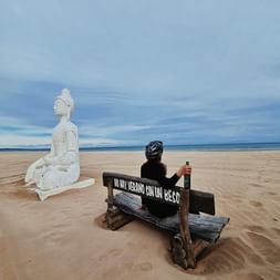Radfahrer mit Helm sitzt auf Holzbank am Sandstrand in Valencia, neben weißer Buddha-Statue. Meer und bewölkter Himmel im Hintergrund.