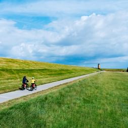 Zwei Radfahrer auf einem asphaltierten Weg durch grüne Deichlandschaft in Pilsum, Ostfriesland. Roter Leuchtturm in der Ferne unter bewölktem Himmel.