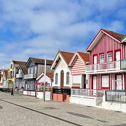 Reihe traditioneller Fischerhäuser in Costa Nova mit bunten vertikalen Streifen in Rot, Grün, Gelb und Weiß entlang einer Kopfsteinpflasterstraße.