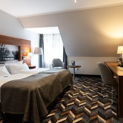 Modernes Doppelzimmer mit weißer Bettwäsche, grauem Überwurf, geometrischem Teppich, Schreibtisch, Sessel und Wandfernseher im Hotel Zollhaus Rastede.