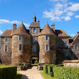 Mittelalterliches Steinschloss in Saint-Sauveur-en-Puisaye mit mehreren Türmen und Kegeldächern, umgeben von geschnittenen Hecken und Steinmauer.