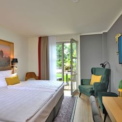 Helles Hotelzimmer mit Doppelbett, gelben Kissen, grünem Sessel, Wandfernseher und Balkontür im Landhotel Schnuck in der Lüneburger Heide.