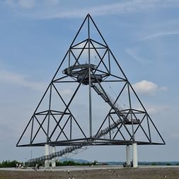 Große Stahlpyramide mit Aussichtsplattform und Wendeltreppe vor blauem Himmel. Das Tetraeder-Wahrzeichen steht auf einem Hügel in Essen.