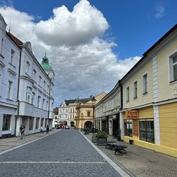 Kopfsteinpflasterstraße in Mělník, gesäumt von bunten historischen Gebäuden unter dramatischem Wolkenhimmel. Weiße, gelbe und beige Fassaden.