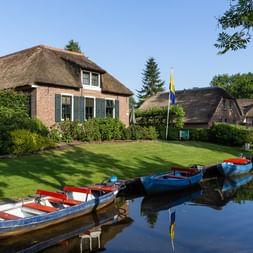 Reihe bunter Boote am Kanal in Giethoorn mit traditionellen Reetdachhäusern, grünen Rasenflächen und Bäumen unter blauem Himmel.