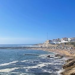 Küstenblick auf Ericeira mit weißen Gebäuden auf Klippen, Sandstrand mit Besuchern, felsiger Küste und blauem Atlantik.