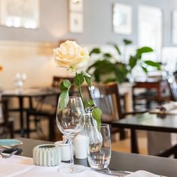 Elegantes Restaurant-Interieur mit weißer Rose in Vase auf gedecktem Tisch. Weingläser und Gedeck sichtbar mit dunklen Holzstühlen im Hintergrund.