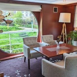 Elegante Bibliothek-Lounge auf der SE Manon mit beigen Sesseln um einen Holztisch, großes Panoramafenster mit Blick auf grüne Uferland.