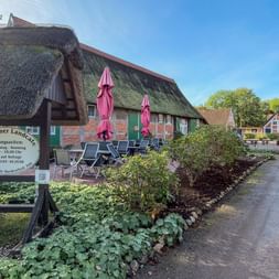 Traditionelles reetgedecktes Café in Mooriem mit Außenbestuhlung, Holzschild und Fachwerkhäusern entlang eines gepflasterten Weges unter blauem Himmel.
